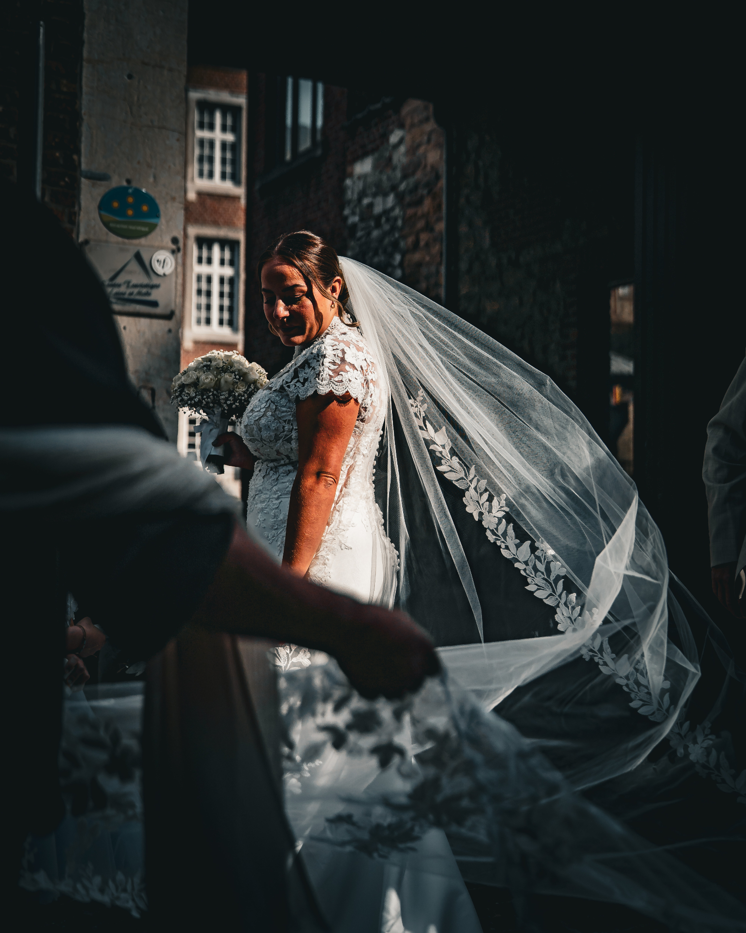 Mariée dans une ruelle, voile dans le vent et bouquet de roses blanches